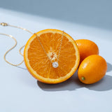 Dainty Solitaire Necklace on a pale blue backdrop beside a halved orange and two kumquats