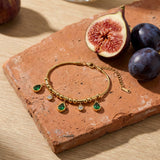 Emerald Teardrop Charm Bracelet on a weathered terracotta tile with a cut fig and dark plums