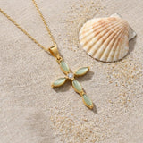 Green Cat's Eye Cross Necklace on raw linen beside a scallop shell and a scatter of fine sand