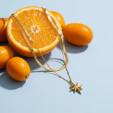 North Star Layered Necklace on a pale blue backdrop beside a halved orange and two kumquats