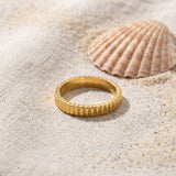 Ribbed Thin Band Ring on raw linen beside a scallop shell and a scatter of fine sand