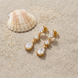 Triple CZ Drop Earrings on raw linen beside a scallop shell and a scatter of fine sand