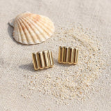 Ridged gold bar stud earrings on raw linen beside a scallop shell and a scatter of fine sand