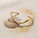Wavy Pear Cuff on raw linen beside a scallop shell and a scatter of fine sand